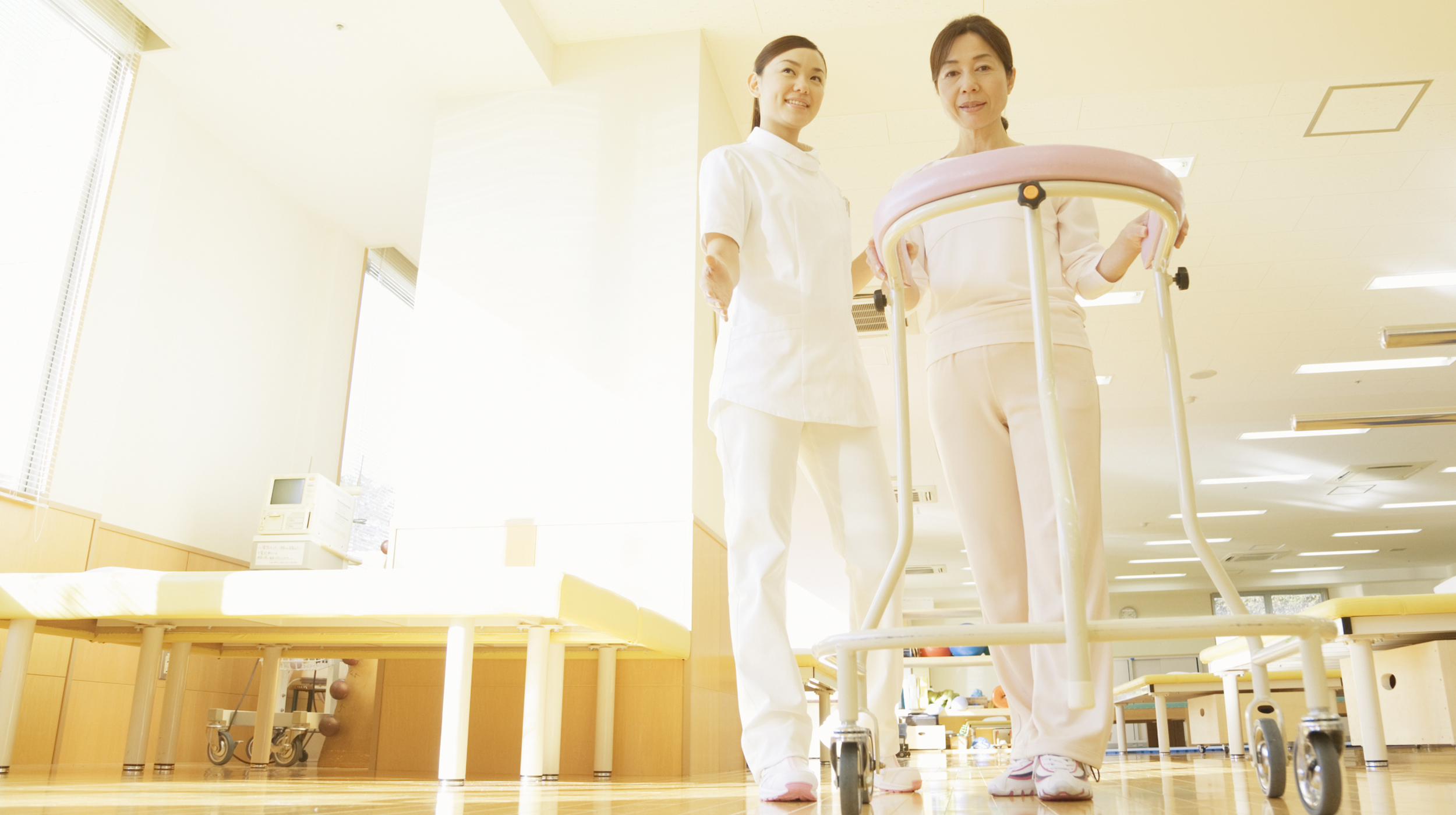 女性の看護師に付き添われて歩行器を使ってリハビリに励む女性の写真